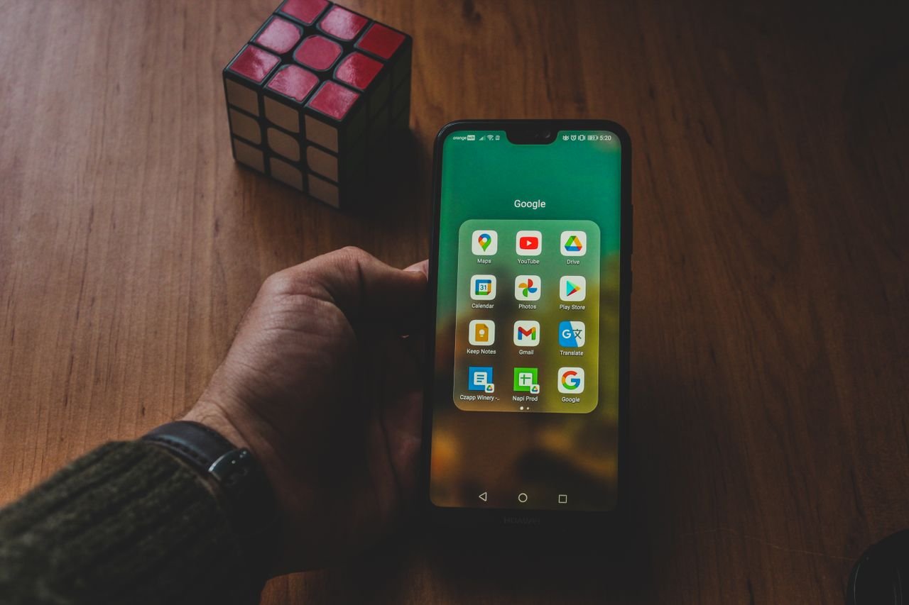 Home A hand holds a smartphone displaying various apps, next to a Rubik's cube on a wooden surface.